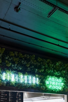 A neon sign displaying 'KIMI SUSHI' and 'ASIAN STREET FOOD' is illuminated against a backdrop of lush green foliage. The dim setting creates a contrast, highlighting the vibrant greens from the sign. The ceiling above features exposed industrial elements.