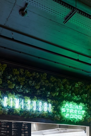 A neon sign displaying 'KIMI SUSHI' and 'ASIAN STREET FOOD' is illuminated against a backdrop of lush green foliage. The dim setting creates a contrast, highlighting the vibrant greens from the sign. The ceiling above features exposed industrial elements.