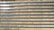 An array of long, parallel rows of solar panels is arranged on a barren, sandy landscape. The panels are aligned neatly, with sunlight reflecting off some of them, creating a shimmering effect. Sparse patches of green are visible between the rows, highlighting the arid environment.