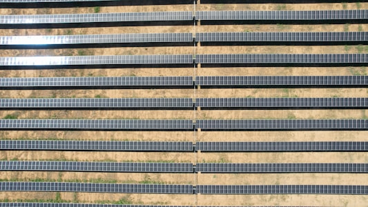 An array of long, parallel rows of solar panels is arranged on a barren, sandy landscape. The panels are aligned neatly, with sunlight reflecting off some of them, creating a shimmering effect. Sparse patches of green are visible between the rows, highlighting the arid environment.