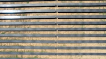 An array of long, parallel rows of solar panels is arranged on a barren, sandy landscape. The panels are aligned neatly, with sunlight reflecting off some of them, creating a shimmering effect. Sparse patches of green are visible between the rows, highlighting the arid environment.