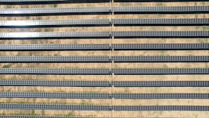 An array of long, parallel rows of solar panels is arranged on a barren, sandy landscape. The panels are aligned neatly, with sunlight reflecting off some of them, creating a shimmering effect. Sparse patches of green are visible between the rows, highlighting the arid environment.