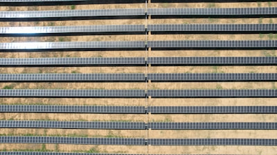 An array of long, parallel rows of solar panels is arranged on a barren, sandy landscape. The panels are aligned neatly, with sunlight reflecting off some of them, creating a shimmering effect. Sparse patches of green are visible between the rows, highlighting the arid environment.