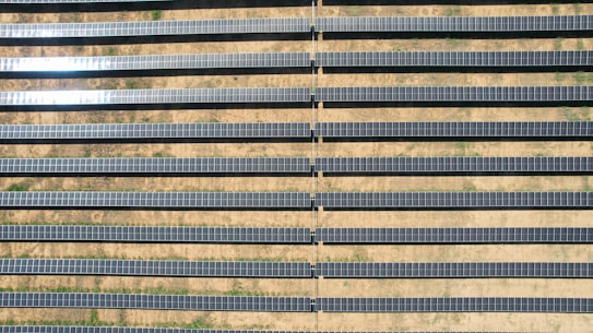 An array of long, parallel rows of solar panels is arranged on a barren, sandy landscape. The panels are aligned neatly, with sunlight reflecting off some of them, creating a shimmering effect. Sparse patches of green are visible between the rows, highlighting the arid environment.