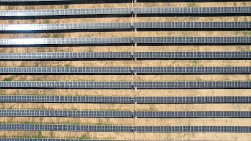 An array of long, parallel rows of solar panels is arranged on a barren, sandy landscape. The panels are aligned neatly, with sunlight reflecting off some of them, creating a shimmering effect. Sparse patches of green are visible between the rows, highlighting the arid environment.