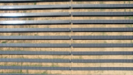 An array of long, parallel rows of solar panels is arranged on a barren, sandy landscape. The panels are aligned neatly, with sunlight reflecting off some of them, creating a shimmering effect. Sparse patches of green are visible between the rows, highlighting the arid environment.