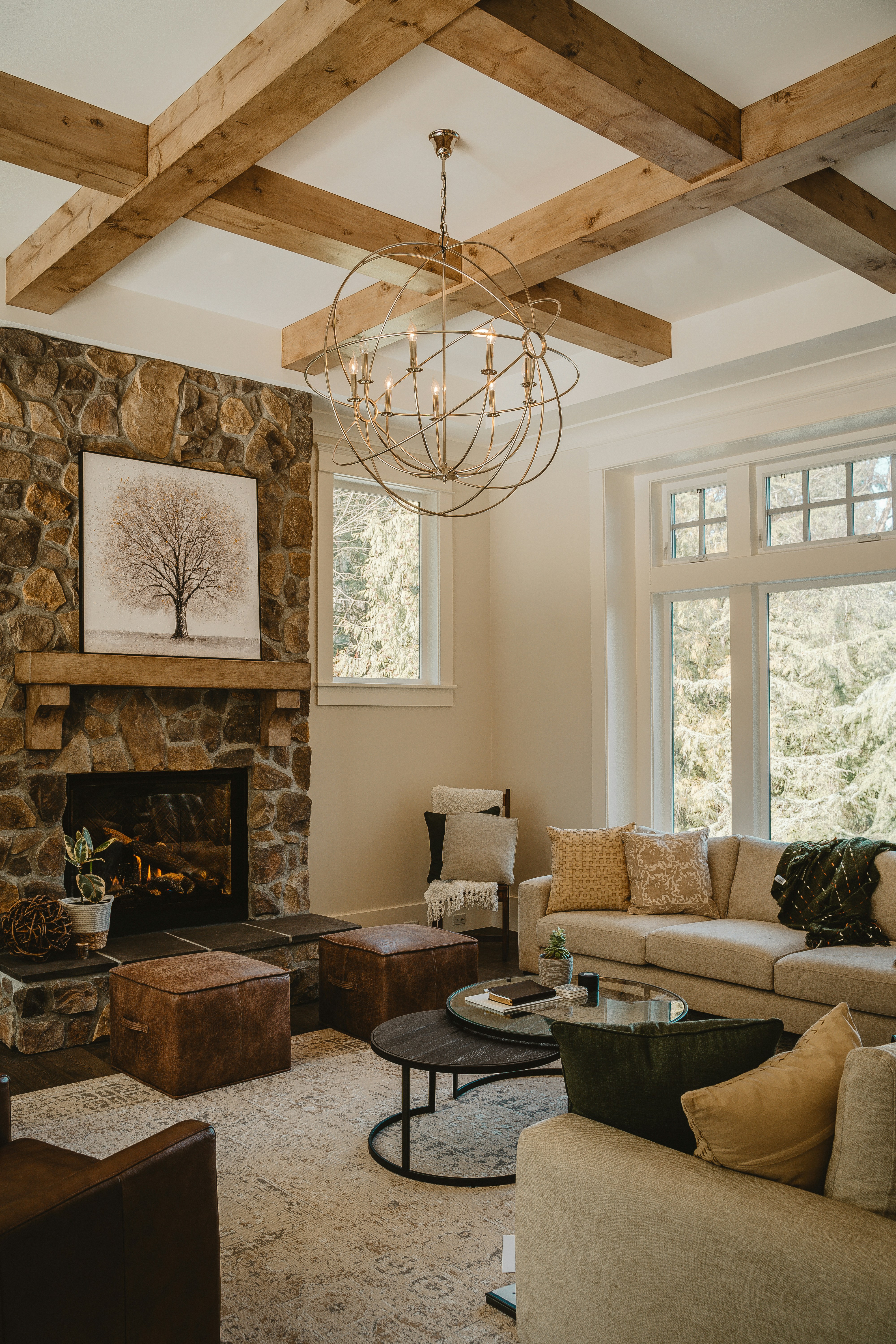 A cozy living room with a stone fireplace and a mantle displaying a tree artwork. The room features a comfortable beige sectional sofa accented with pillows, a large window offering natural light, and a stylish round coffee table. The ceiling showcases wooden beams and a decorative chandelier, while plush ottomans and a patterned rug complete the warm setting.