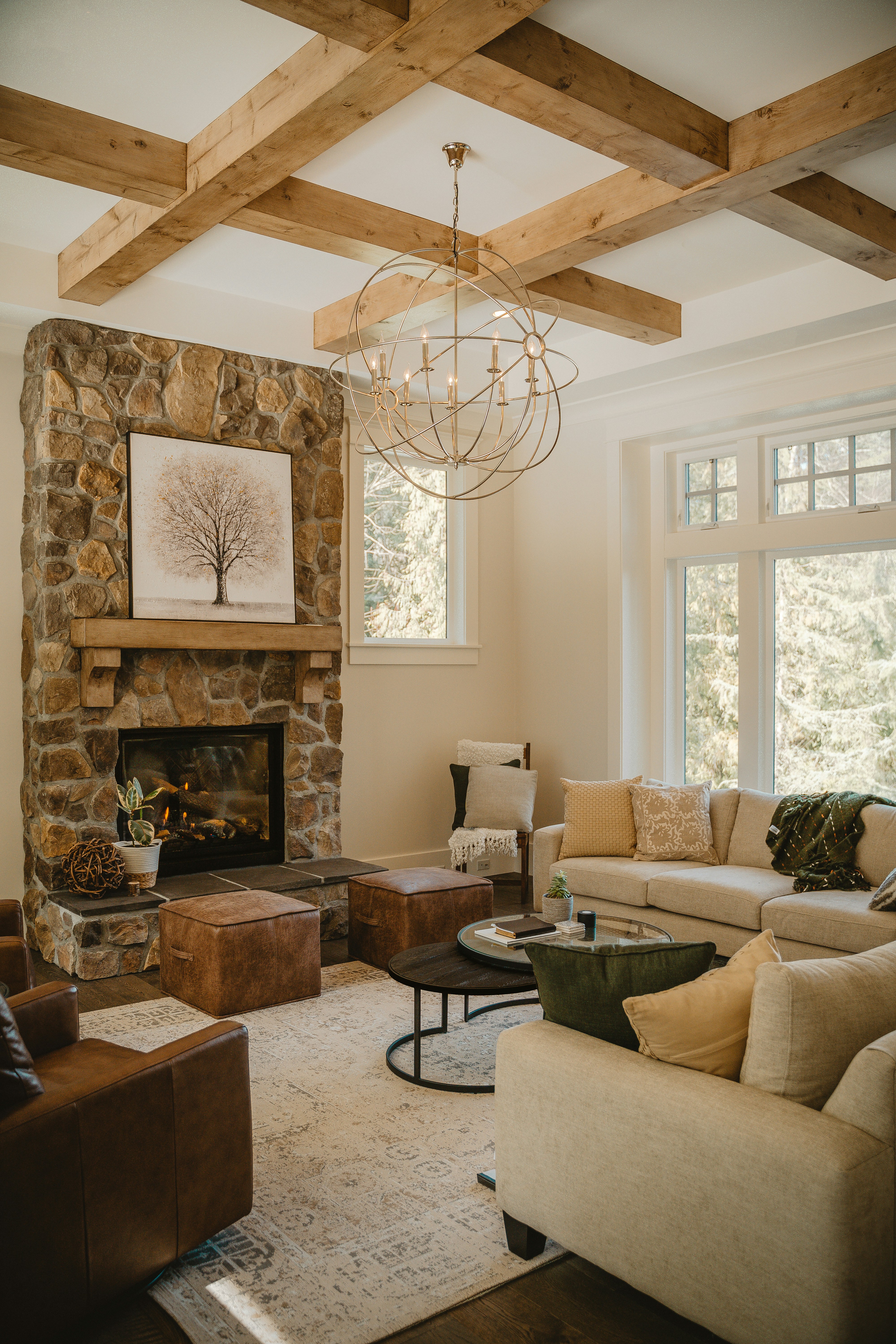 A cozy living room with a stone fireplace as the focal point. The room features a modern chandelier hanging from a ceiling with exposed wooden beams. Comfortable seating includes a beige sectional sofa and leather armchairs. Earthy tones are prevalent, with wooden and leather elements complementing the decor. A large window allows natural light to enter, brightening the space. The room is decorated with plants and a framed artwork above the fireplace that depicts a tree.