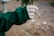 A close-up of a hand wearing a green sleeve holds a rolled cigarette or joint. The background is a blurred outdoor setting that suggests a natural, wooded area.
