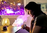 A person is sitting at a table, exhaling vapor from what appears to be a vape device. The room is dimly lit, with a warm lamp on the table casting a soft yellow glow. Behind the person, a window shows bright purple and pink city lights, adding a colorful backdrop.