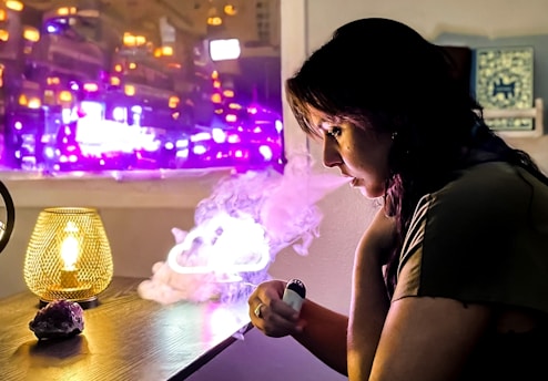 A person is sitting at a table, exhaling vapor from what appears to be a vape device. The room is dimly lit, with a warm lamp on the table casting a soft yellow glow. Behind the person, a window shows bright purple and pink city lights, adding a colorful backdrop.