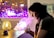 A person is sitting at a table, exhaling vapor from what appears to be a vape device. The room is dimly lit, with a warm lamp on the table casting a soft yellow glow. Behind the person, a window shows bright purple and pink city lights, adding a colorful backdrop.