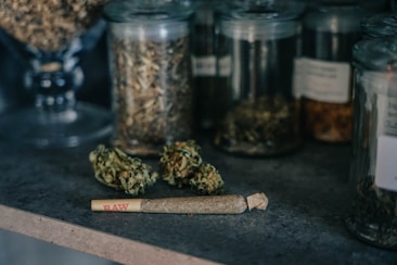Several glass jars containing dried herbs and plant material are arranged on a countertop. In front of the jars, there is a rolled cigarette or joint labeled 'RAW' lying next to some loose herbs or cannabis buds.