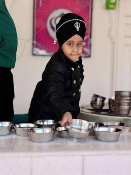 A child wearing a black turban adorned with a decorative emblem and beads is standing in front of a table with multiple stainless steel bowls. The child is dressed in a black jacket and appears to be in a candid moment, looking directly ahead. The background includes a purple and white poster and a partial view of another person wearing a green garment.