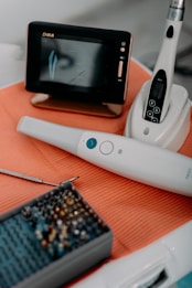 A dental setup is displayed with various tools and equipment. A monitor is showing a dental graphic, alongside tools like a scaler and a set of dental burs. The items are laid out on an orange mat.
