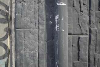A textured wall surface with a vertical pipe running through it. The wall appears weathered, with cracked and peeling paint. The surface has a slightly embossed pattern, and there is a faded graffiti design partially visible on the left side.