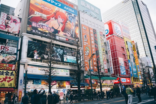 A busy urban street filled with colorful billboards and advertisements featuring anime characters and vibrant graphics. The street is lined with multi-story buildings showcasing various shops and businesses. People are walking along the sidewalk, some stopping to take photos or look at the signs. The scene is lively and bustling, with a mix of cultural and commercial elements.