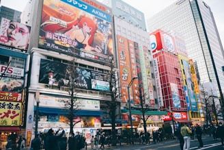 A busy urban street filled with colorful billboards and advertisements featuring anime characters and vibrant graphics. The street is lined with multi-story buildings showcasing various shops and businesses. People are walking along the sidewalk, some stopping to take photos or look at the signs. The scene is lively and bustling, with a mix of cultural and commercial elements.