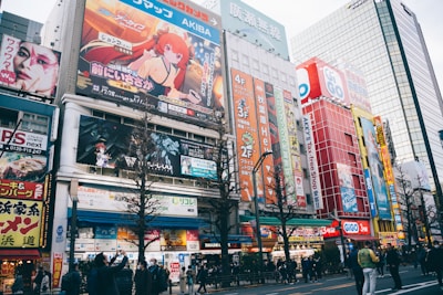 A busy urban street filled with colorful billboards and advertisements featuring anime characters and vibrant graphics. The street is lined with multi-story buildings showcasing various shops and businesses. People are walking along the sidewalk, some stopping to take photos or look at the signs. The scene is lively and bustling, with a mix of cultural and commercial elements.
