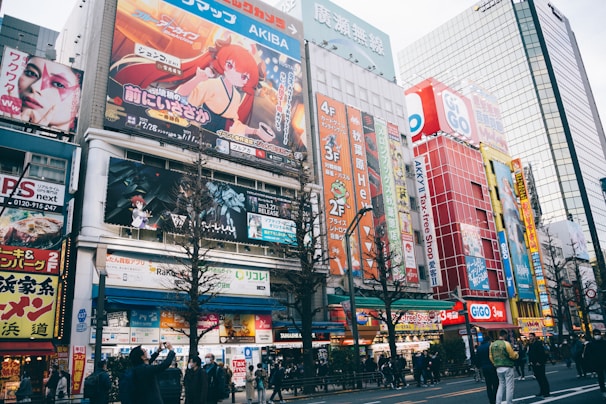 A busy urban street filled with colorful billboards and advertisements featuring anime characters and vibrant graphics. The street is lined with multi-story buildings showcasing various shops and businesses. People are walking along the sidewalk, some stopping to take photos or look at the signs. The scene is lively and bustling, with a mix of cultural and commercial elements.