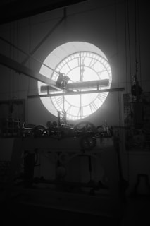 A large, intricate clock mechanism stands against the backdrop of a circular window with Roman numerals that let in natural light. The room appears filled with various mechanical parts and industrial elements. The clock parts are a combination of gears, pulleys, and levers.