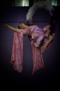 A person performs an aerial act using a hoop adorned with feathers. They wear a flowing, soft pink outfit that drapes elegantly as they hang gracefully. The dim lighting adds a theatrical atmosphere to the scene.