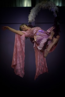 A person performs an aerial act using a hoop adorned with feathers. They wear a flowing, soft pink outfit that drapes elegantly as they hang gracefully. The dim lighting adds a theatrical atmosphere to the scene.