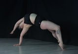 A person performs a dynamic bridge pose against a dark background, wearing black athletic wear. The lighting highlights the contours of the body, emphasizing the strength and flexibility required for this pose. A tattoo is visible on one of the legs. The floor is a light color, contrasting with the dark backdrop.