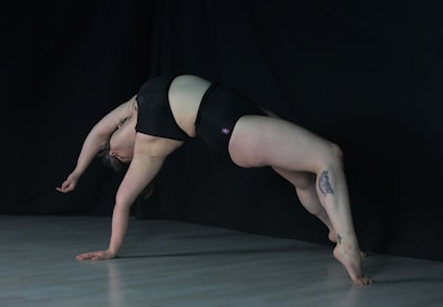 A person performs a dynamic bridge pose against a dark background, wearing black athletic wear. The lighting highlights the contours of the body, emphasizing the strength and flexibility required for this pose. A tattoo is visible on one of the legs. The floor is a light color, contrasting with the dark backdrop.