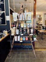 A rustic display inside a cafe featuring a variety of coffee and tea products. Two wooden poles form a triangular frame in front of shelves stacked with different brands and flavors of coffee and tea in bags and boxes. A small potted plant is placed to the side. The setting includes wooden and metal textures with soft lighting, and there is a menu board on the wall at the left.