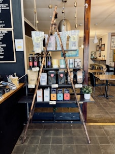 A rustic display inside a cafe featuring a variety of coffee and tea products. Two wooden poles form a triangular frame in front of shelves stacked with different brands and flavors of coffee and tea in bags and boxes. A small potted plant is placed to the side. The setting includes wooden and metal textures with soft lighting, and there is a menu board on the wall at the left.