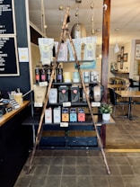 A rustic display inside a cafe featuring a variety of coffee and tea products. Two wooden poles form a triangular frame in front of shelves stacked with different brands and flavors of coffee and tea in bags and boxes. A small potted plant is placed to the side. The setting includes wooden and metal textures with soft lighting, and there is a menu board on the wall at the left.
