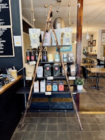 A rustic display inside a cafe featuring a variety of coffee and tea products. Two wooden poles form a triangular frame in front of shelves stacked with different brands and flavors of coffee and tea in bags and boxes. A small potted plant is placed to the side. The setting includes wooden and metal textures with soft lighting, and there is a menu board on the wall at the left.