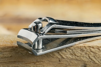 A close-up view of a metallic nail clipper resting on a textured wooden surface. The shiny metal reflects light, highlighting its smooth and polished finish.