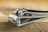 A close-up view of a metallic nail clipper resting on a textured wooden surface. The shiny metal reflects light, highlighting its smooth and polished finish.