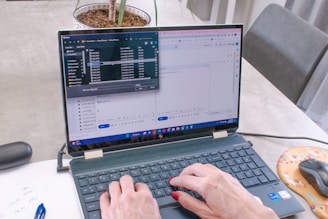 A person with painted nails is typing on a laptop that displays an open file explorer and a Google form in split-screen mode. A pen, a piece of paper with some notes, a mouse on an orange mousepad, and a plant in a pot are visible on the table.