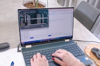 A person with painted nails is typing on a laptop that displays an open file explorer and a Google form in split-screen mode. A pen, a piece of paper with some notes, a mouse on an orange mousepad, and a plant in a pot are visible on the table.