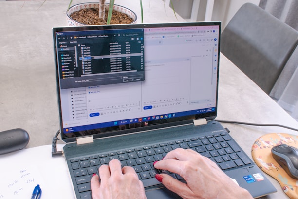 A person with painted nails is typing on a laptop that displays an open file explorer and a Google form in split-screen mode. A pen, a piece of paper with some notes, a mouse on an orange mousepad, and a plant in a pot are visible on the table.