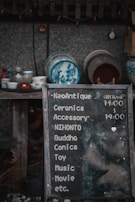 A weathered chalkboard sign lists various items such as ceramics, accessories, comics, and more, with hours indicated. Behind it, a collection of traditional ceramic dishes with blue designs is displayed on wooden shelves against a textured wall.
