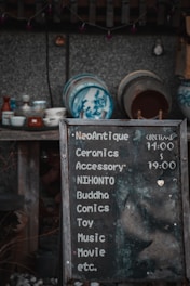 A weathered chalkboard sign lists various items such as ceramics, accessories, comics, and more, with hours indicated. Behind it, a collection of traditional ceramic dishes with blue designs is displayed on wooden shelves against a textured wall.