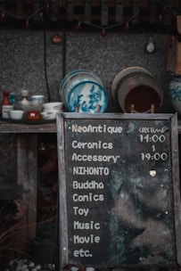 A weathered chalkboard sign lists various items such as ceramics, accessories, comics, and more, with hours indicated. Behind it, a collection of traditional ceramic dishes with blue designs is displayed on wooden shelves against a textured wall.