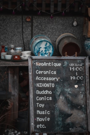 A weathered chalkboard sign lists various items such as ceramics, accessories, comics, and more, with hours indicated. Behind it, a collection of traditional ceramic dishes with blue designs is displayed on wooden shelves against a textured wall.