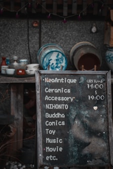 A weathered chalkboard sign lists various items such as ceramics, accessories, comics, and more, with hours indicated. Behind it, a collection of traditional ceramic dishes with blue designs is displayed on wooden shelves against a textured wall.
