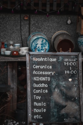 A weathered chalkboard sign lists various items such as ceramics, accessories, comics, and more, with hours indicated. Behind it, a collection of traditional ceramic dishes with blue designs is displayed on wooden shelves against a textured wall.