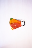 A colorful face mask with a tie-dye pattern, featuring vibrant shades of orange, red, yellow, and green. The mask has black elastic ear loops and is placed against a plain white background, highlighting its vivid design.