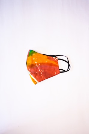 A colorful face mask with a tie-dye pattern, featuring vibrant shades of orange, red, yellow, and green. The mask has black elastic ear loops and is placed against a plain white background, highlighting its vivid design.