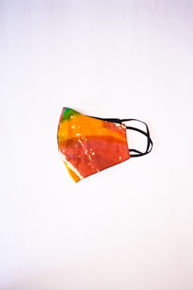 A colorful face mask with a tie-dye pattern, featuring vibrant shades of orange, red, yellow, and green. The mask has black elastic ear loops and is placed against a plain white background, highlighting its vivid design.