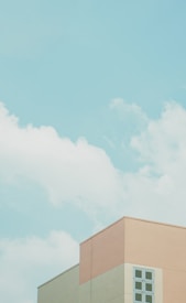 A pastel-toned building facade with a minimalist design sits against a backdrop of a clear blue sky with scattered white clouds.