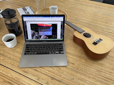 A workspace setup on a wooden surface with a laptop displaying a web page, a ukulele, a French press with coffee, and two cups. There is also a glass container and some documents on the side.