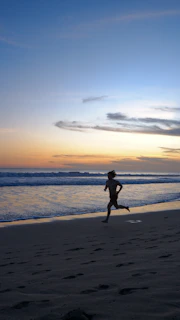 A runner sprinting along a sunlit sandy beach with waves gently crashing nearby.
